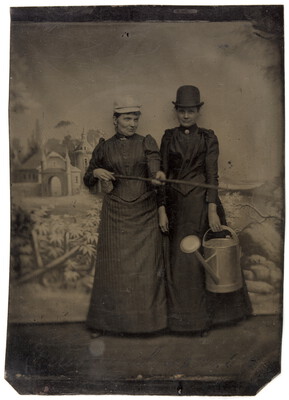 Zwei Frauen posieren mit Haushalts- oder Gartenrequisiten vor gemaltem Hintergrund